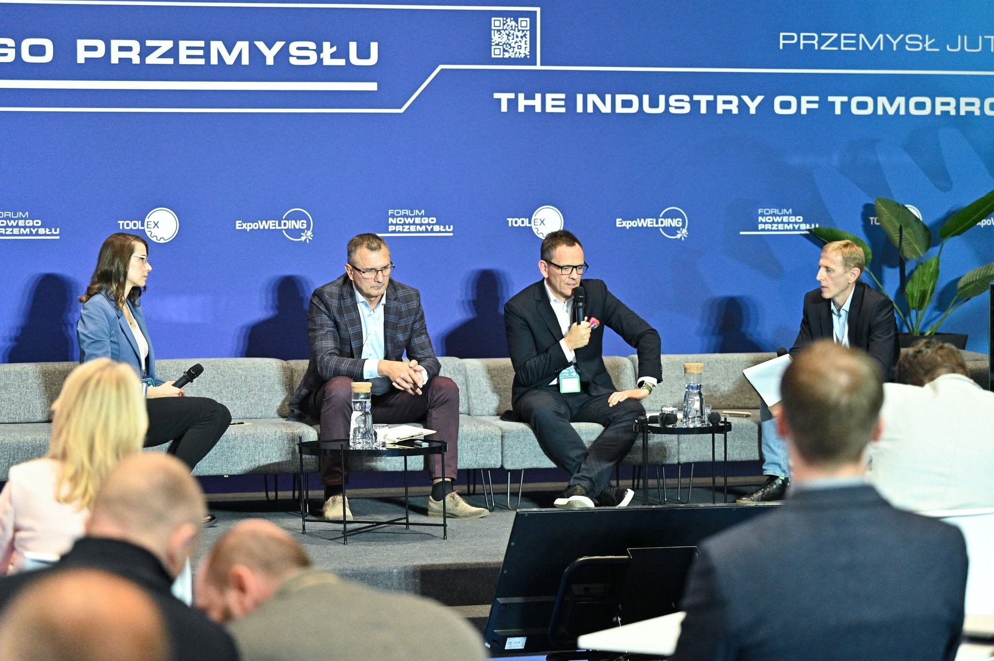 Na zdjęciu widać fotele z trzema prelegentami i jedną prelegentką. na widowni widoczni ludzie